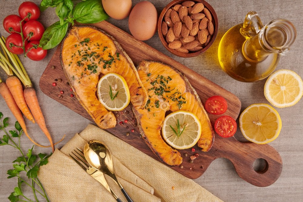 Due bistecche di salmone al forno con limone su un tagliere, accanto a una bottiglia d'olio, mandorle, uova, pomodori e carote