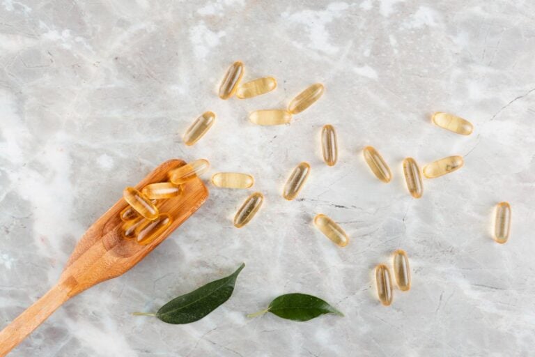 Capsule di omega-3 su marmo chiaro, alcune su un cucchiaio di legno, accanto a due foglie verdi